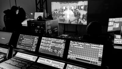 Black and white photo of a backstage production control room with mixing boards and monitors showing a live fashion runway