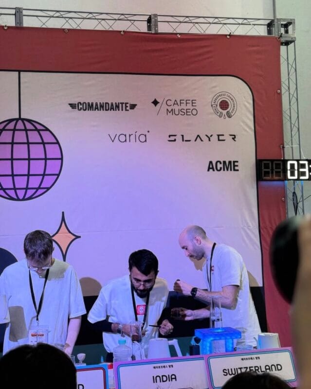 Competitors from India and Switzerland brewing at the World AeroPress Championship stage, sponsor banners behind them