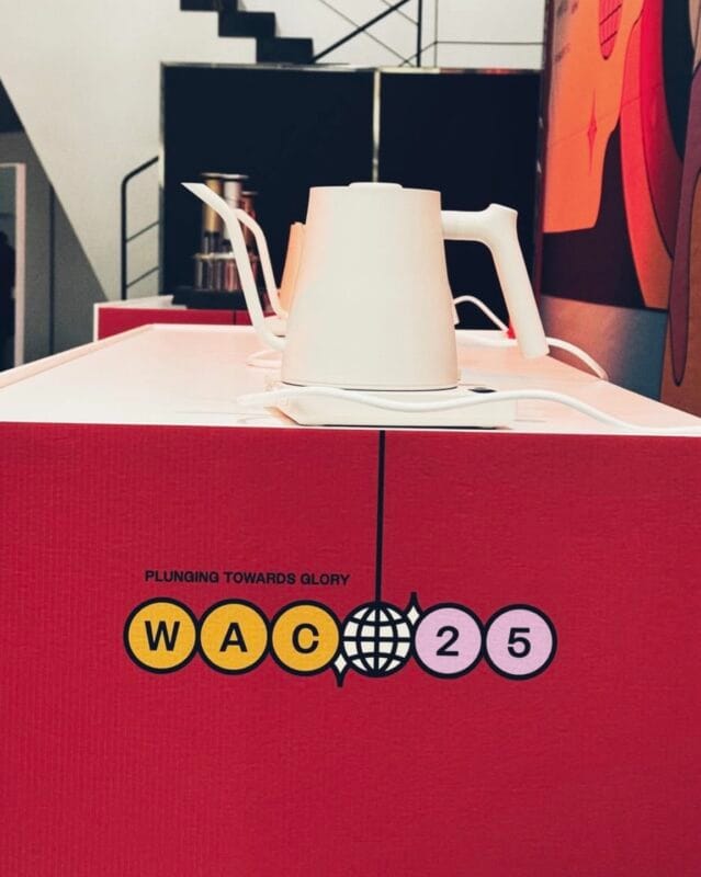 White electric gooseneck kettle on a red WAC 25 branded station reading Plunging Towards Glory, with stairs and orange garment visible in the background