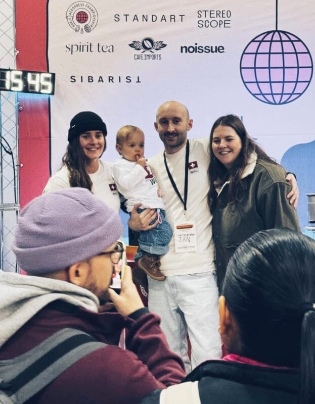 Competitor Jan from Switzerland holding a baby and posing with two smiling women in front of a World Aeropress Championship sponsor banner