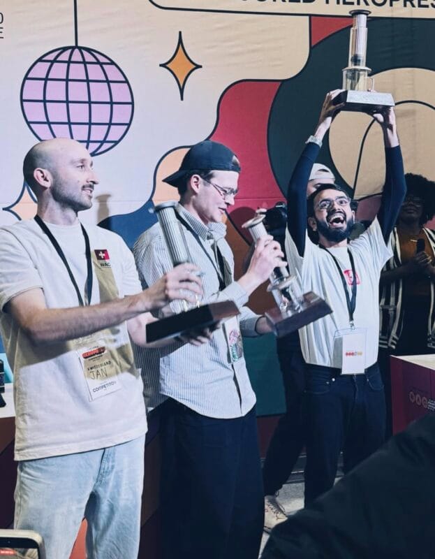 Three competitors raising their Aeropress trophies on stage at the World Aeropress Championship, the rightmost competitor lifting his trophy high overhead