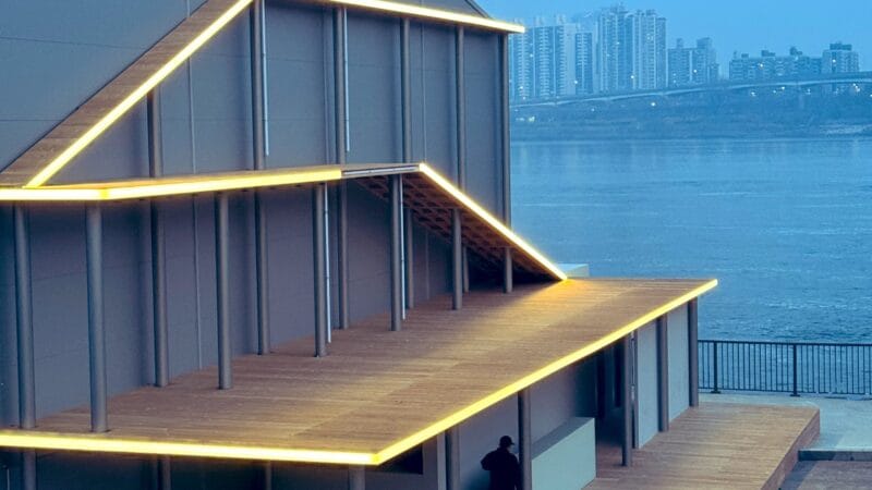 Illuminated multi-level wooden pavilion beside a river at dusk, city skyline visible in the background