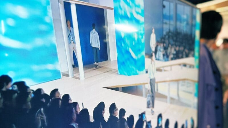 Fashion show audience watching a model walk a runway framed by large LED video walls displaying ocean imagery