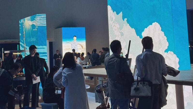 Fashion show venue interior with guests mingling near tall LED video walls showing beach and sky imagery