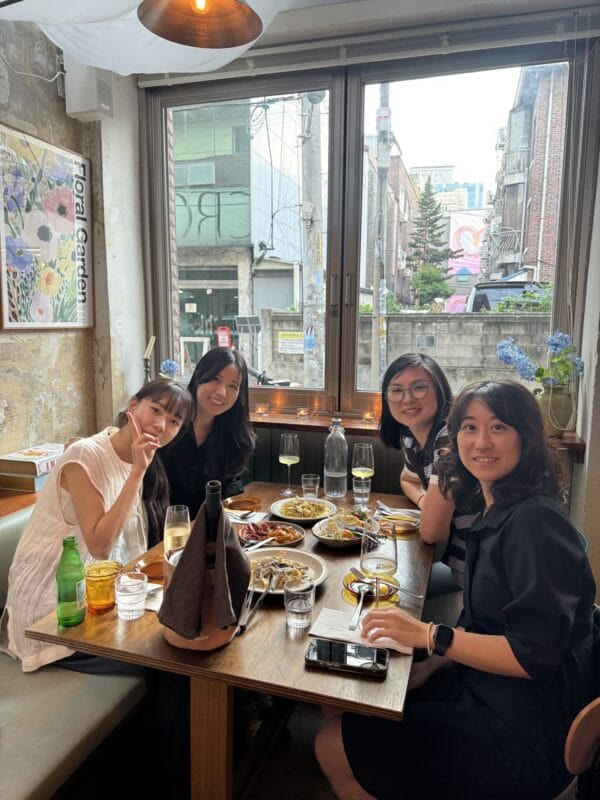 Four women seated at a restaurant table sharing plates of food, smiling at the camera